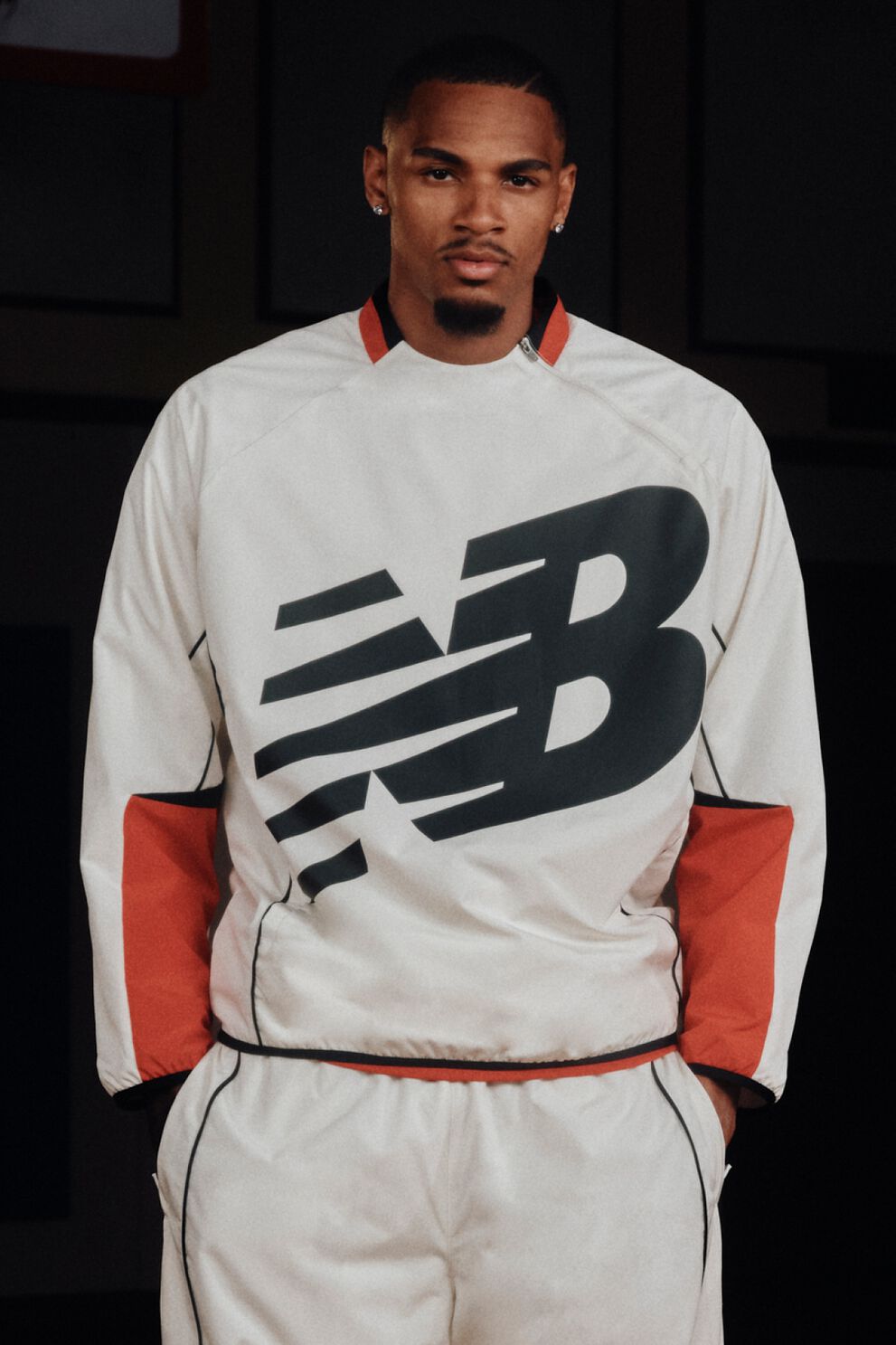 Dejounte Murray wearing a white New Balance basketball warm-up top with a large black logo, standing on an indoor basketball court.