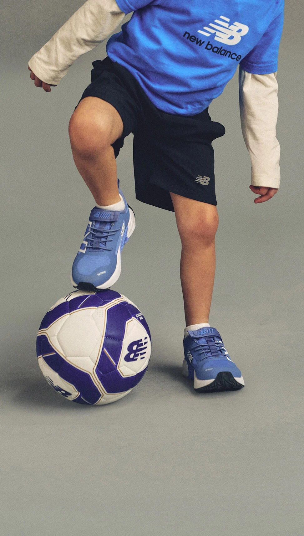 Child wearing a blue New Balance shirt, black shorts, and blue sneakers, balancing one foot on a purple-and-white soccer ball against a plain background.