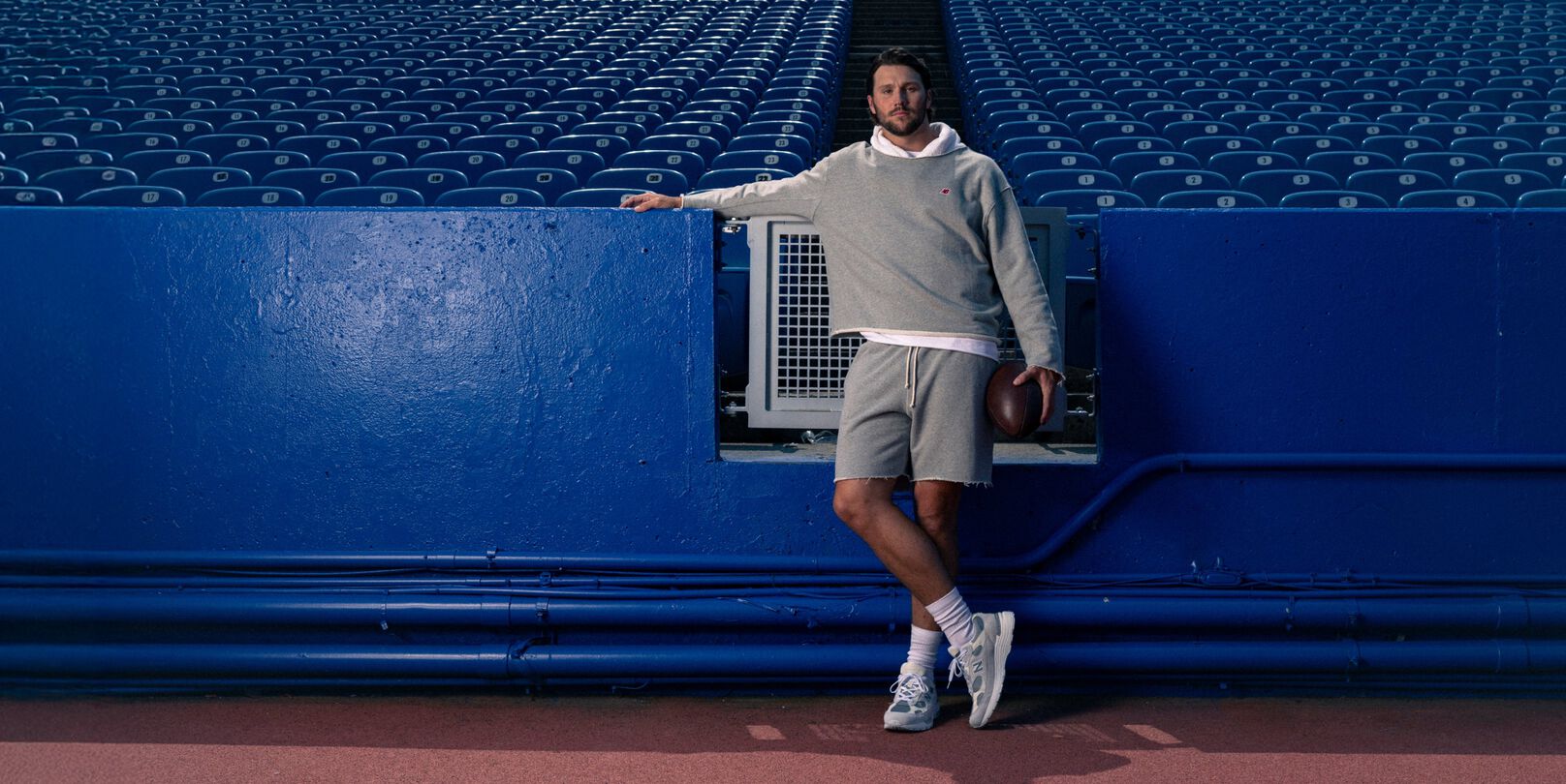 Young male athlete in a grey hoodie leaning on a stadium fence.
