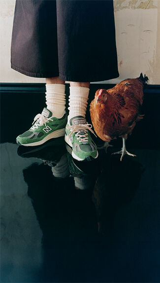 Knee down view of a man wearing New Balance Made in UK sneakers standing next to a chicken in a room with white walls and a shiny black floor.  