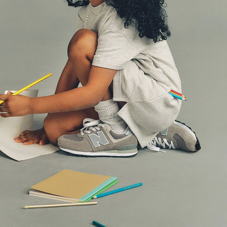 Close-up studio image of a child crouching on the floor against a plain light gray background, focused on drawing on a sheet of lined paper. 