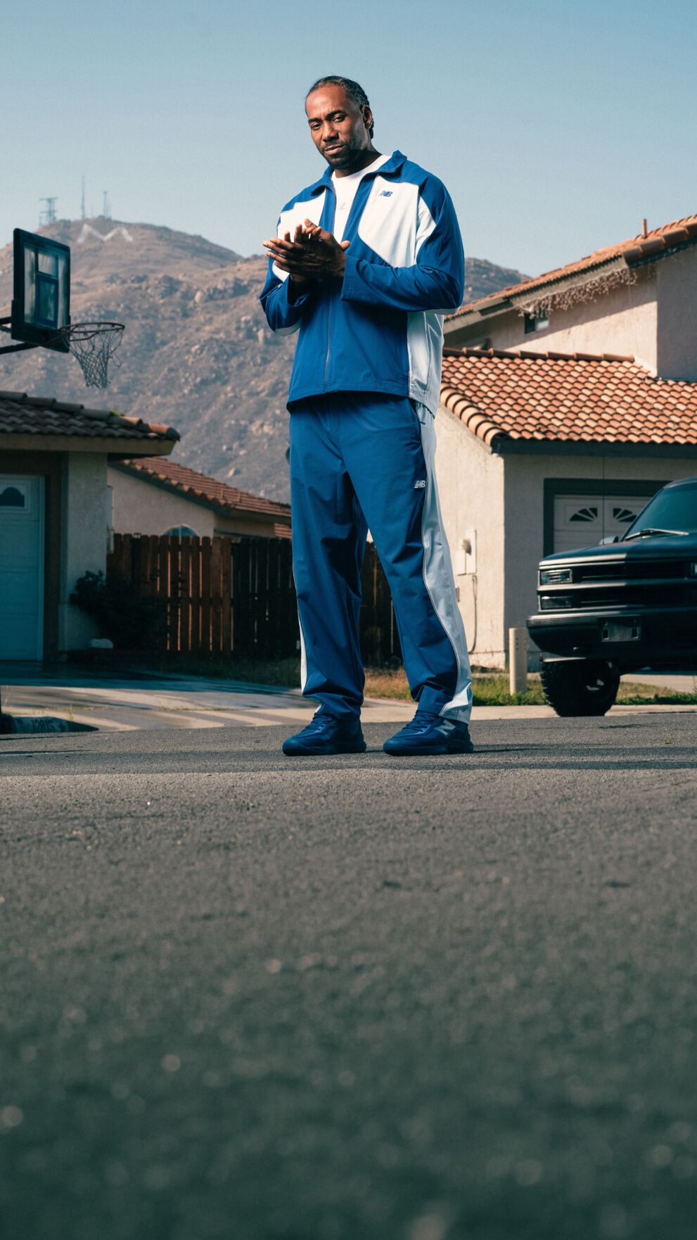 Person wearing a blue and white tracksuit and matching sneakers stands on a suburban street with mountains and houses in the background.