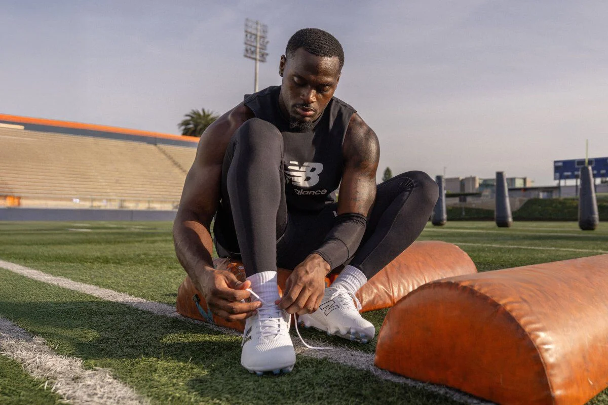 Football athlete in black training gear sits on a football field tying white cleats, with orange padded training equipment nearby.