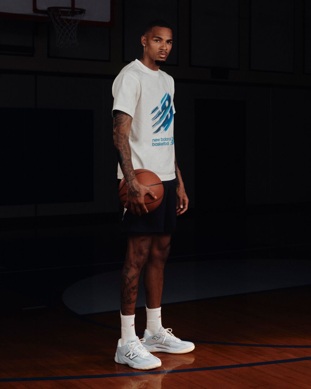 Person standing on an indoor basketball court holding a basketball while wearing a white New Balance Basketball graphic T-shirt, black shorts, and light blue sneakers.