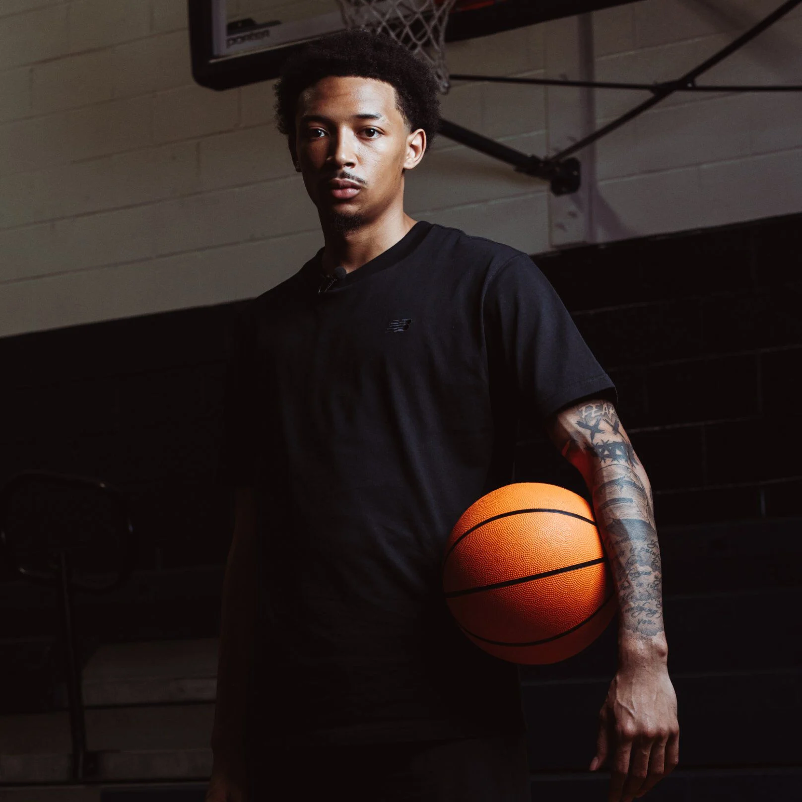A person stands on an indoor basketball court wearing a black T‑shirt and holding a basketball against their hip.