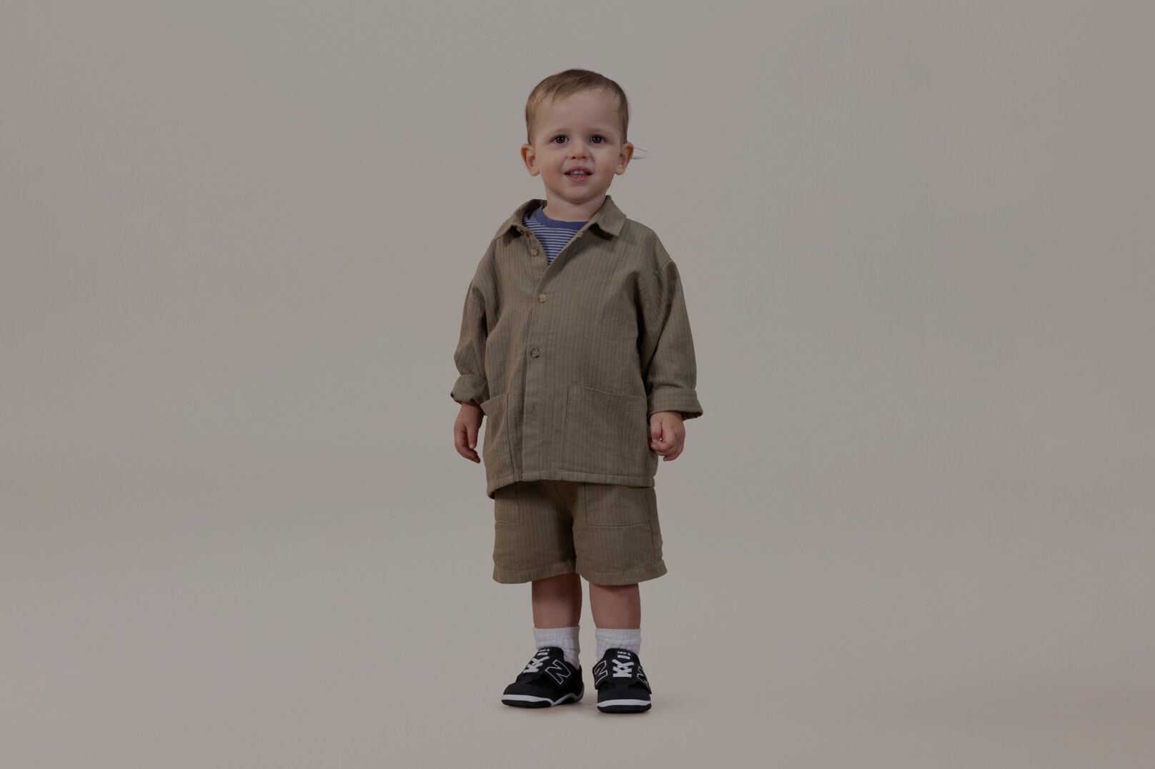 Bebé con un traje color canela de pie sobre un fondo gris.
