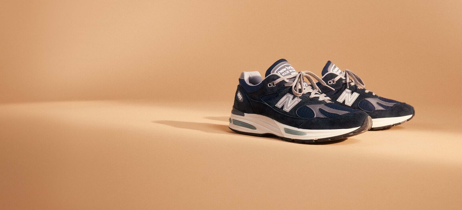 A pair of MADE in UK 991v2 in navy colorway with a tan background.