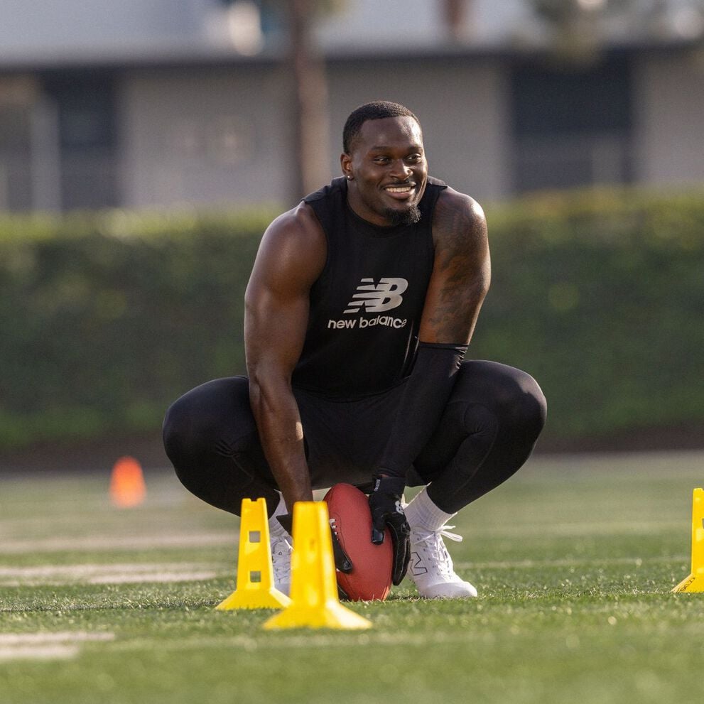Young man crouches on a grass field holding a football between yellow cones during an agility setup, wearing black training apparel and white cleats.