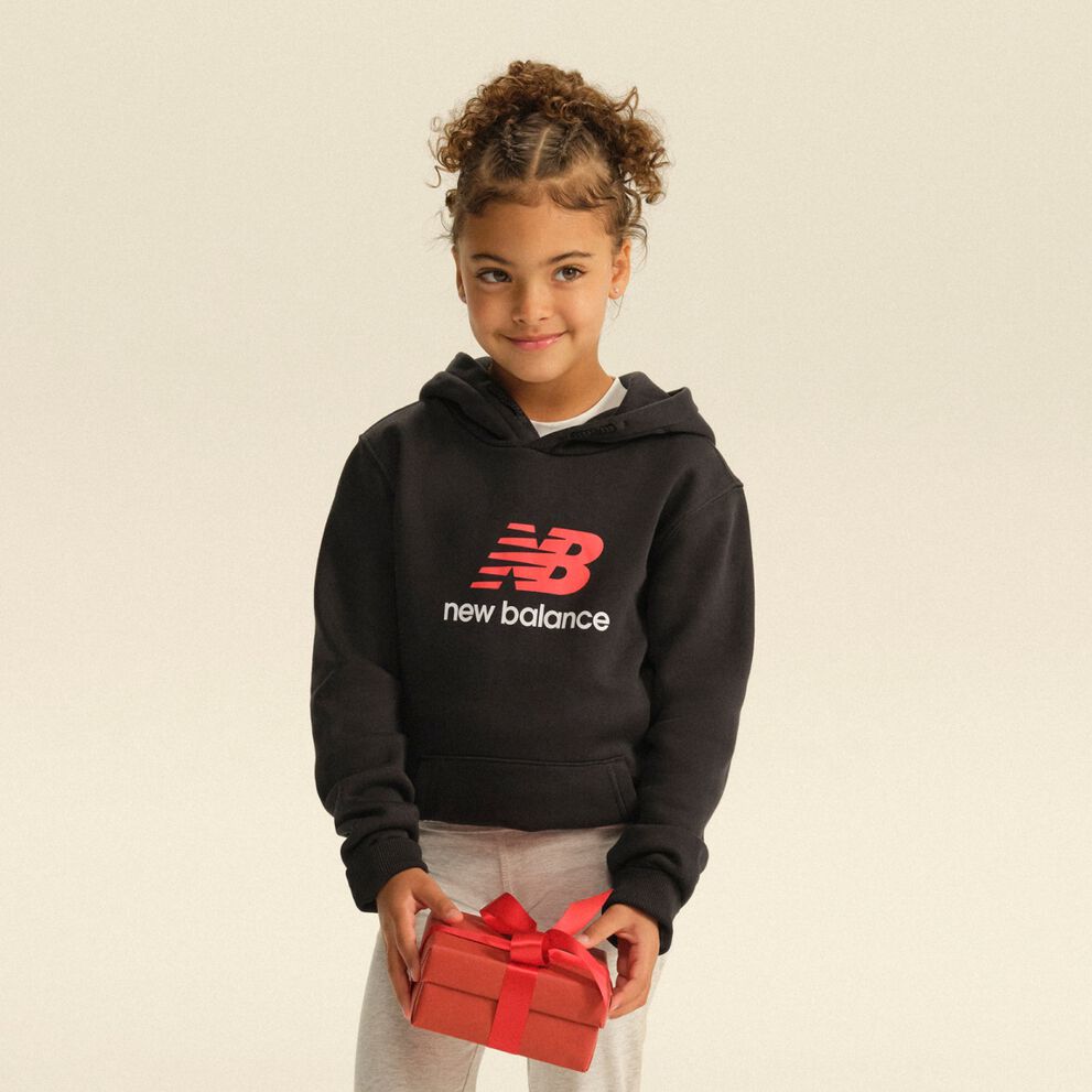 Child wearing a black New Balance hoodie and gray sweatpants while holding a red gift box in a clean studio setting.