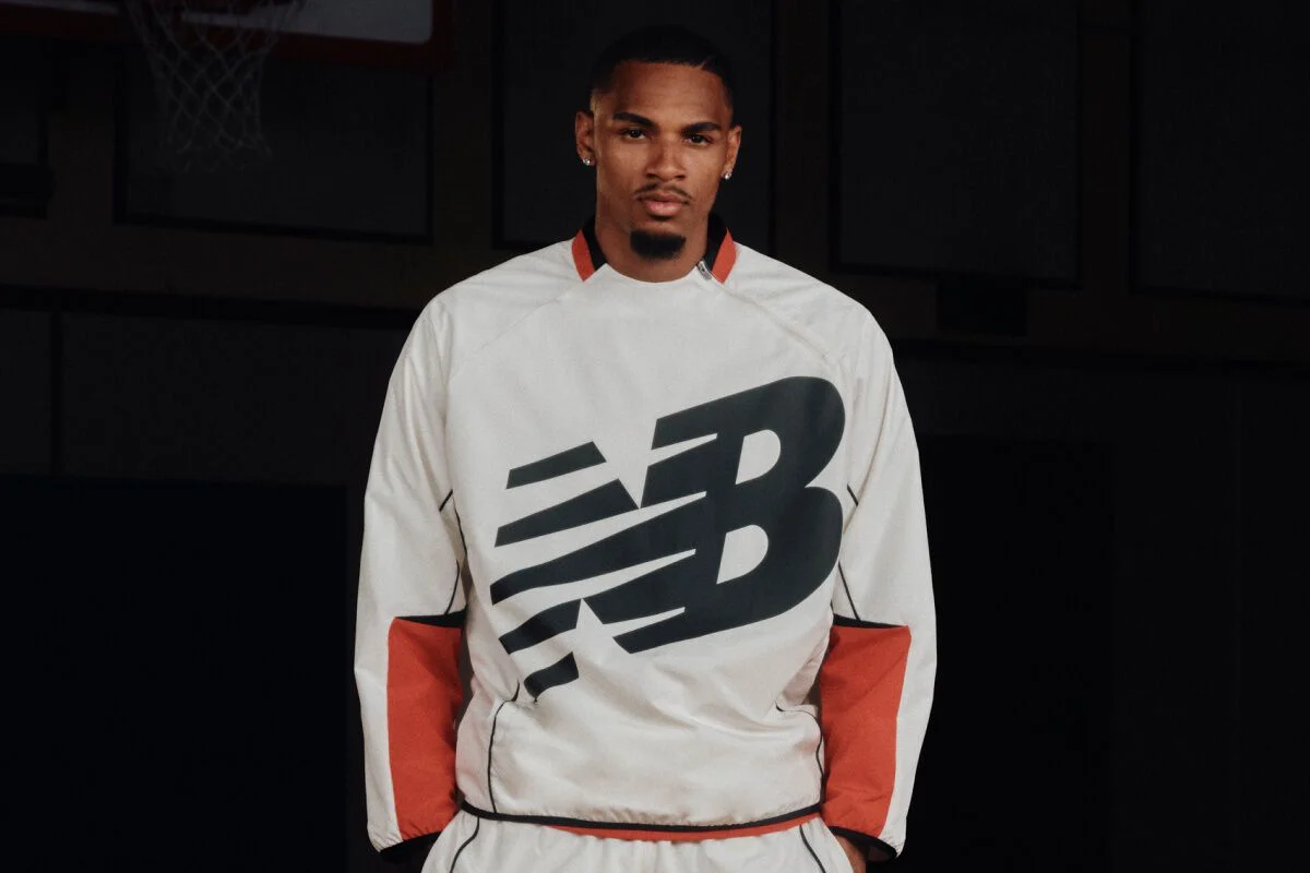 Dejounte Murray wearing a white New Balance basketball warm-up top with a large black logo, standing on an indoor basketball court.