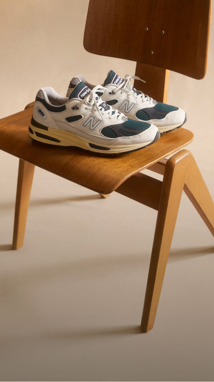 A pair of New Balance Made in UK sneakers sitting on a wooden chair in a beige studio environment. 