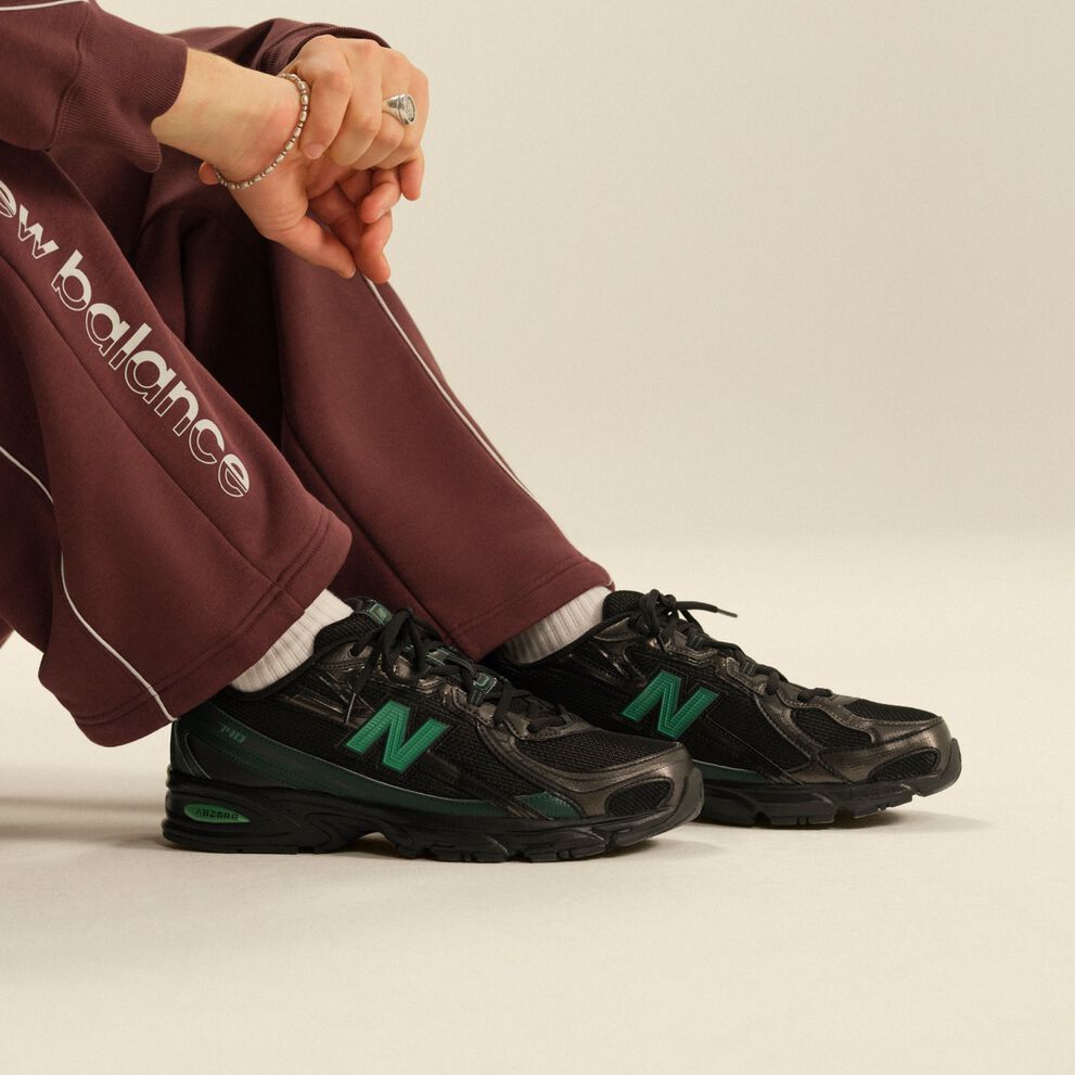Close-up of black New Balance athletic sneakers with green accents paired with burgundy track pants and white crew socks in a clean studio setting.