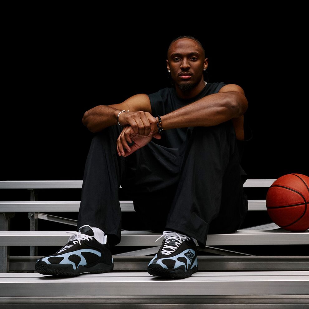 Person seated on metal bleachers beside a basketball, highlighting black-and-light-blue athletic sneakers in a dark studio setting.