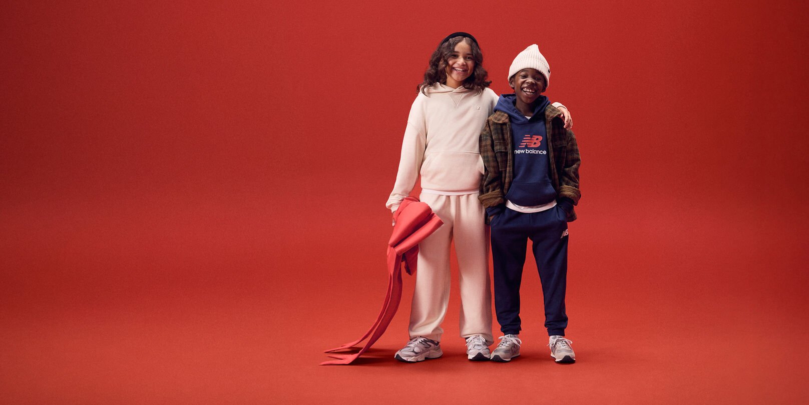 Two young kids smiling with arms around each other on a red background.