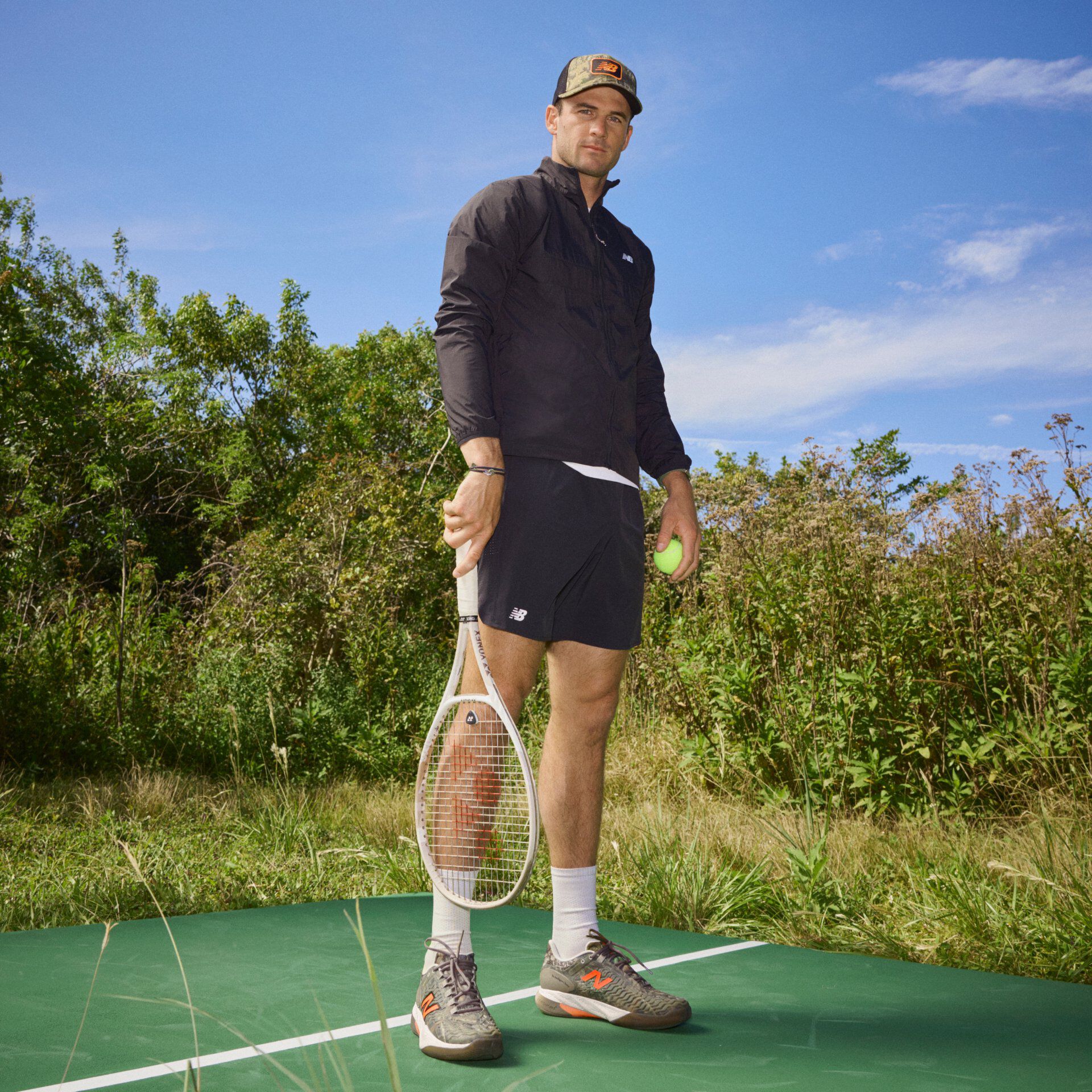 Tommy Paul standing on a tennis court holding a racket, dressed in a black New Balance jacket, black New Balance shorts, and wearing Camo colored shoes and hat with orange accents.