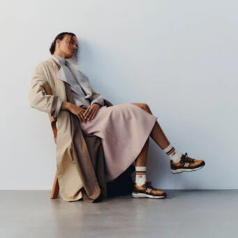 A woman sitting on a wooden chair with her legs crossed wearing a pair of New Balance Made in UK sneakers in a minimalist room with a white wall and concrete floor.