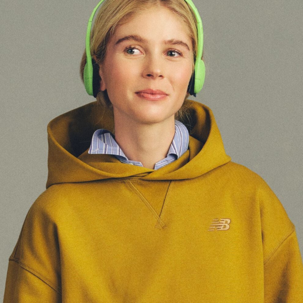 Close-up studio image of a person wearing a mustard-yellow hoodie with a small New Balance logo on the chest, layered over a striped collared shirt.