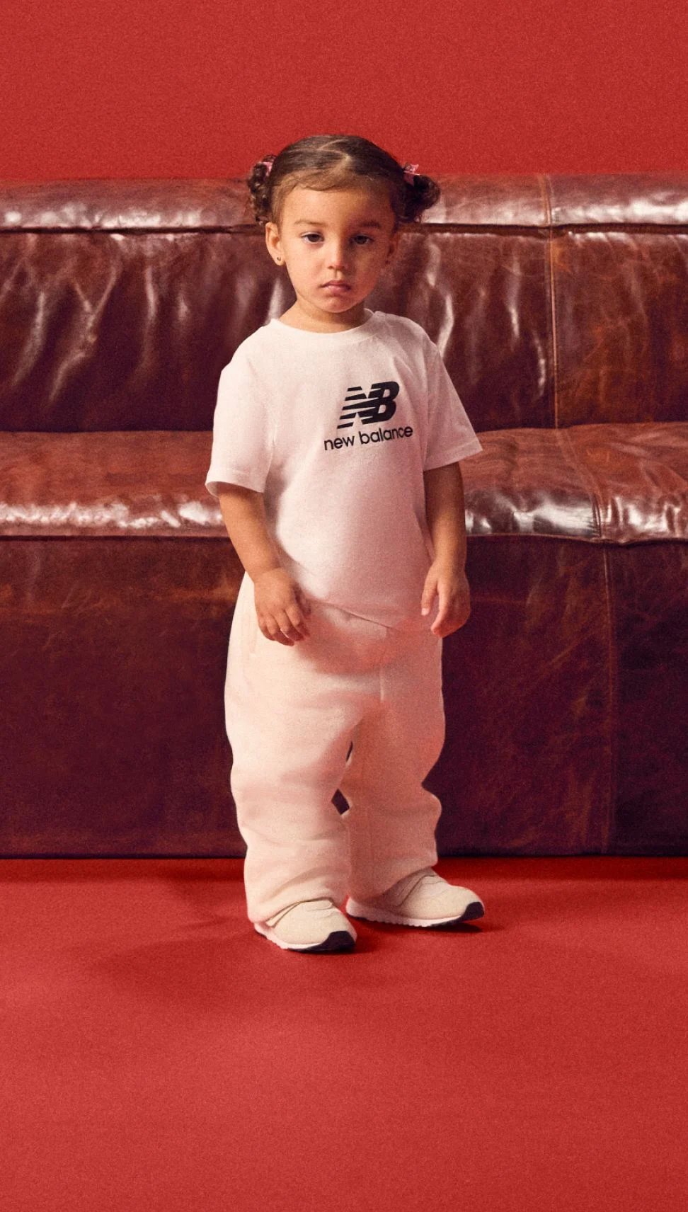 Infant in a white outfit standing in front of a red background. 