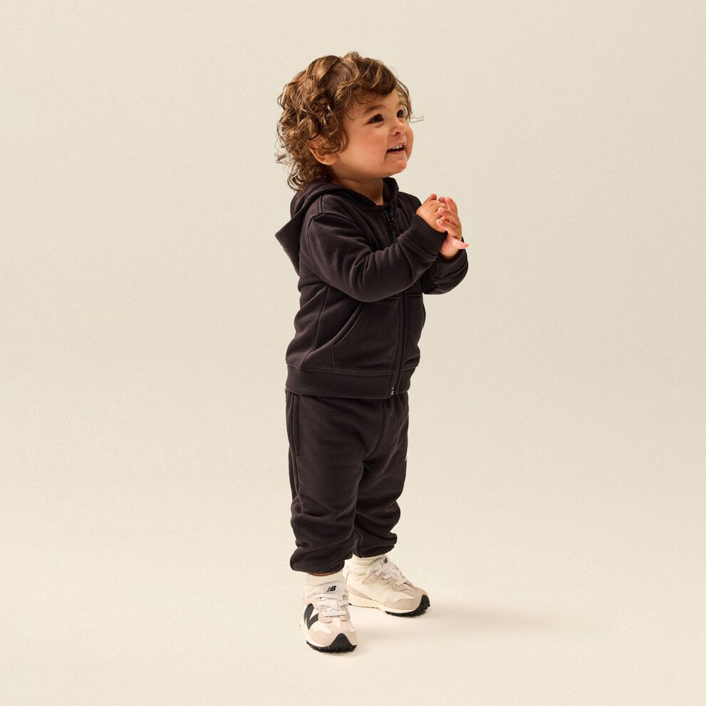 Toddler wearing a black hooded sweatsuit and white New Balance sneakers standing in a clean studio setting.