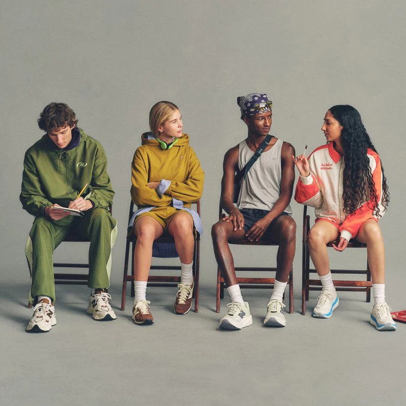Four people seated in a row on chairs against a plain background, each wearing casual outfits and New Balance sneakers, with one writing in a notebook and others conversing.