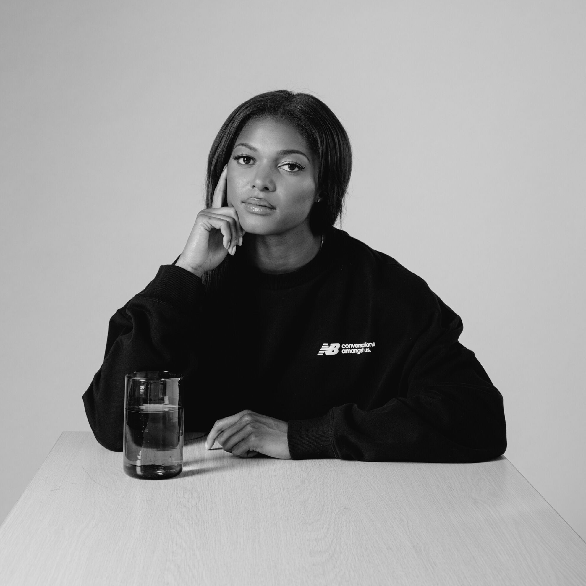 Image Alt Text Black and white image of Gabby Thomas sitting at a table with a glass of water, wearing a black CAU hoodie