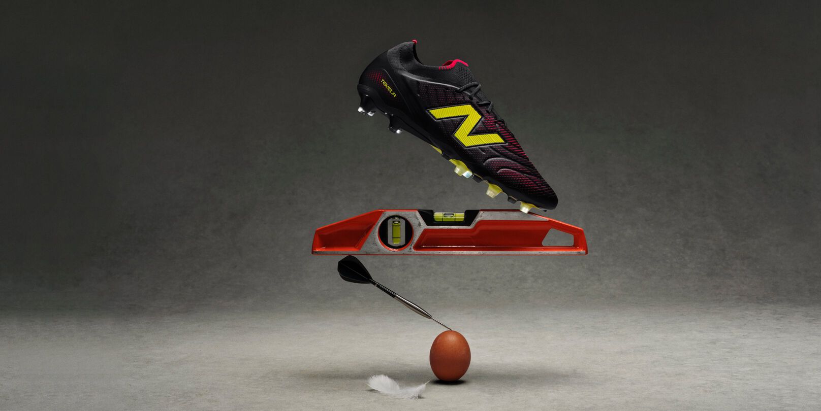 A black soccer cleat with yellow and red accents hovers above an orange leveling tool, which is balanced on a dart tip resting on a brown egg.