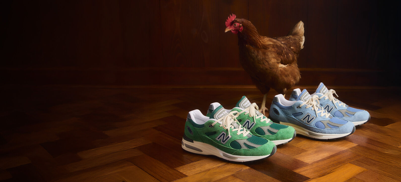 Two pairs of New Balance Made in UK sneakers sitting on a rustic herringbone wooden floor with a chicken standing behind them.  