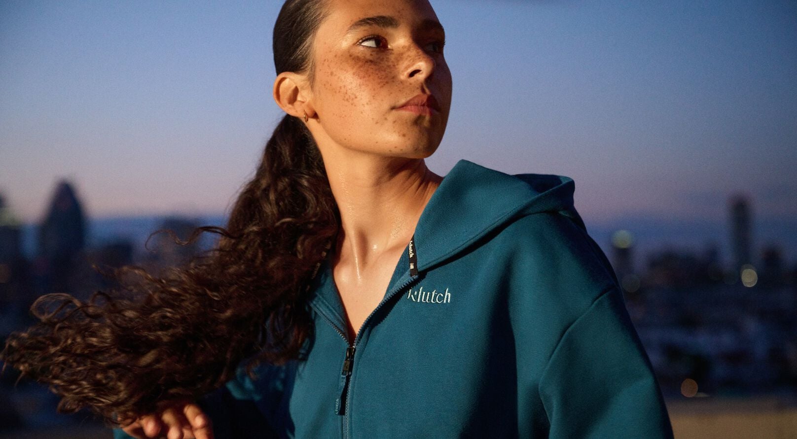 Athlete wearing a teal zip-up hoodie with a city skyline blurred in the background at dusk.