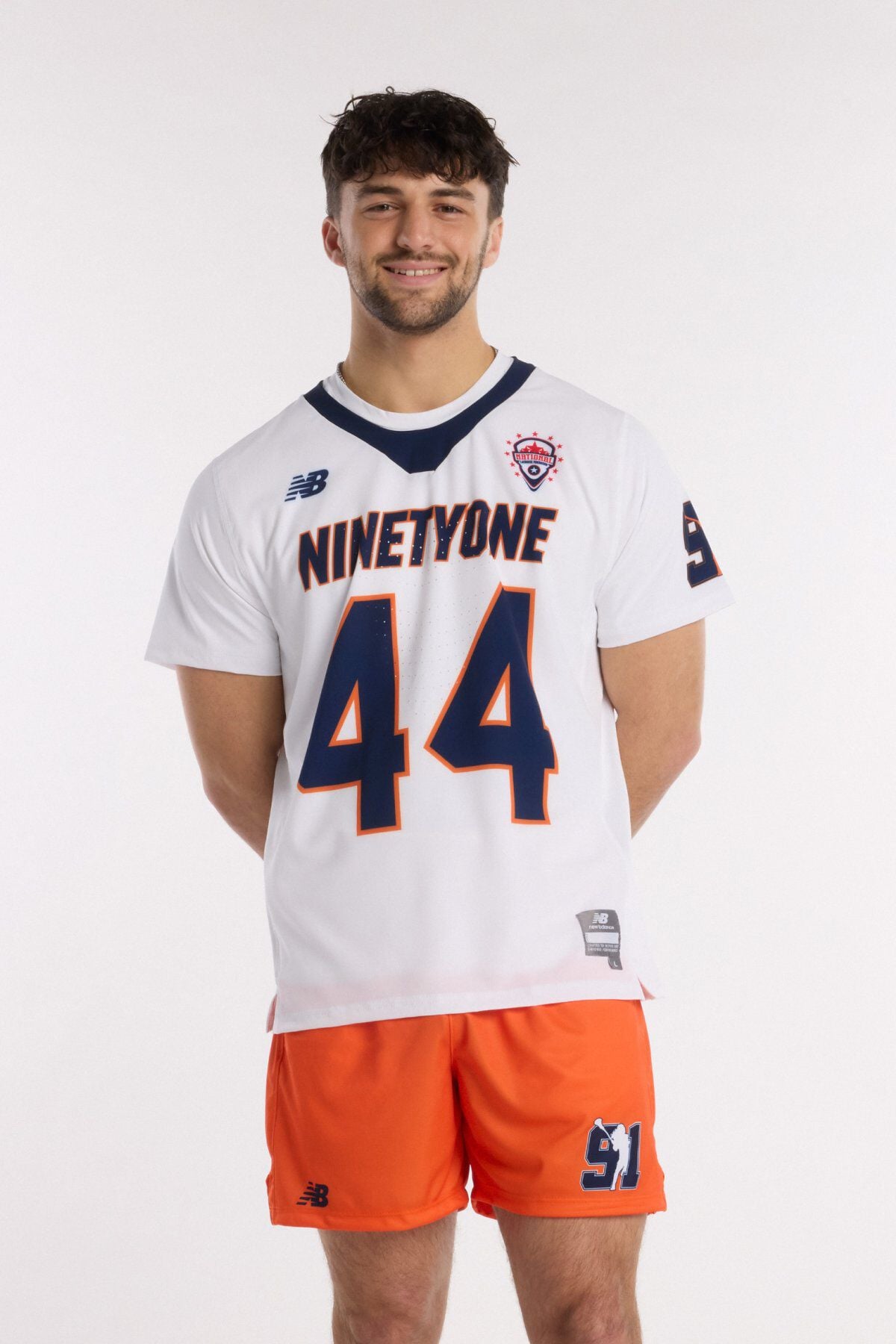 Athlete wearing a white lacrosse jersey with &ldquo;NINETYONE&rdquo; and number 44, paired with orange shorts, standing front-facing.