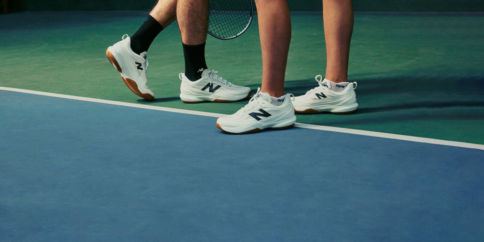 Primer plano de dos tenistas con zapatillas blancas de pista de pie sobre una pista cubierta con una raqueta visible al fondo.