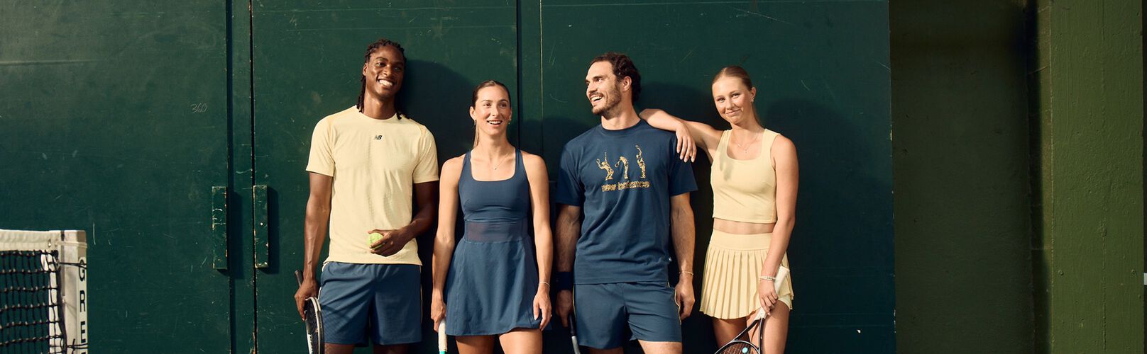 Four tennis players in coordinated athletic apparel stand against a dark green court wall holding rackets and a tennis ball.