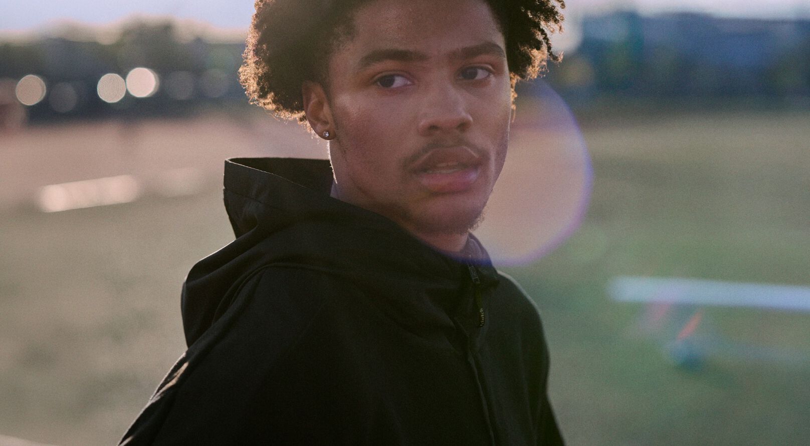 Athlete wearing a black hoodie standing on an outdoor track and field at dusk.