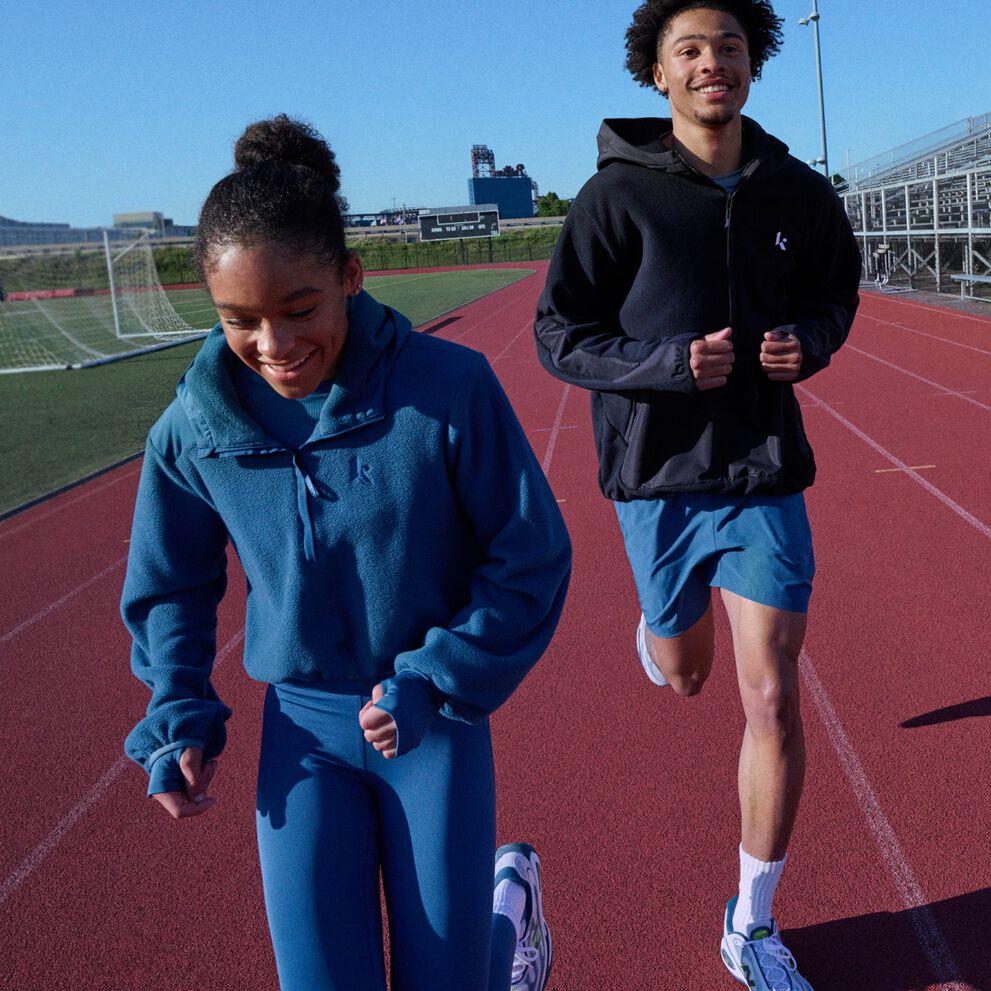 Two athletes running on a track beside stadium bleachers while wearing coordinated athletic apparel.