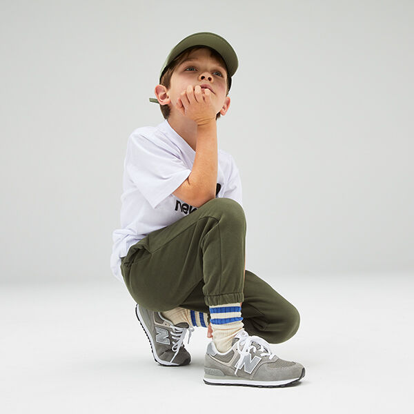 A young boy, crouched in front of a light grey studio backdrop, wearing a white NB Essentials tee, olive green joggers, and grey NB Classic 574 sneakers 