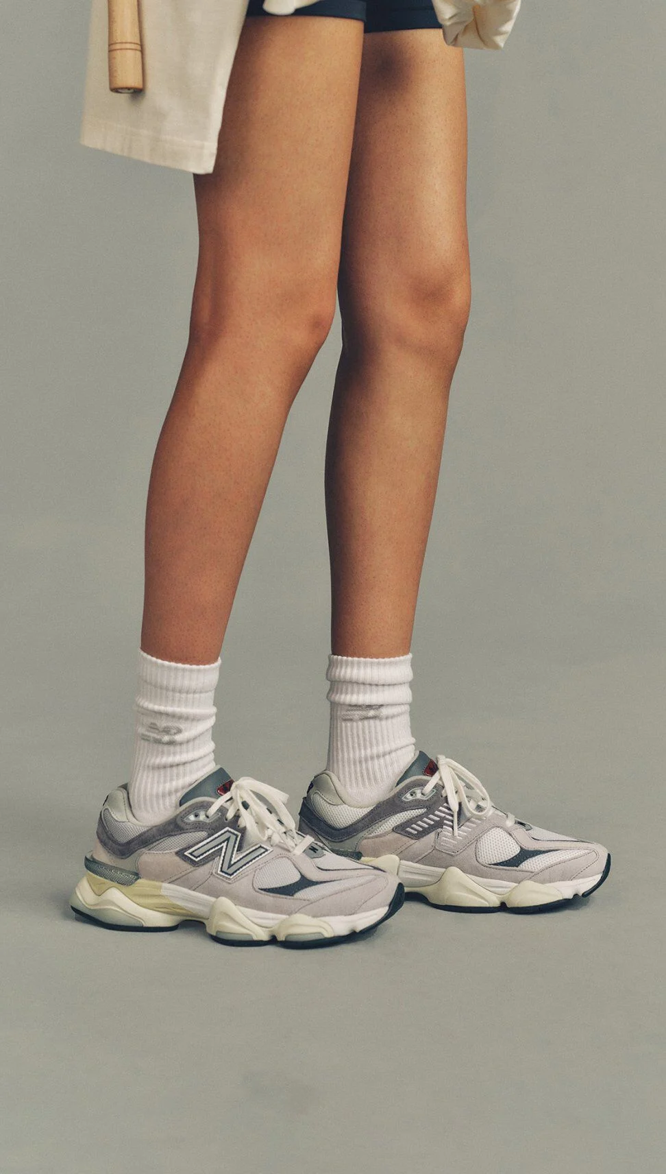Close-up of a person standing in white crew socks and gray New Balance sneakers with layered soles, set against a neutral background.