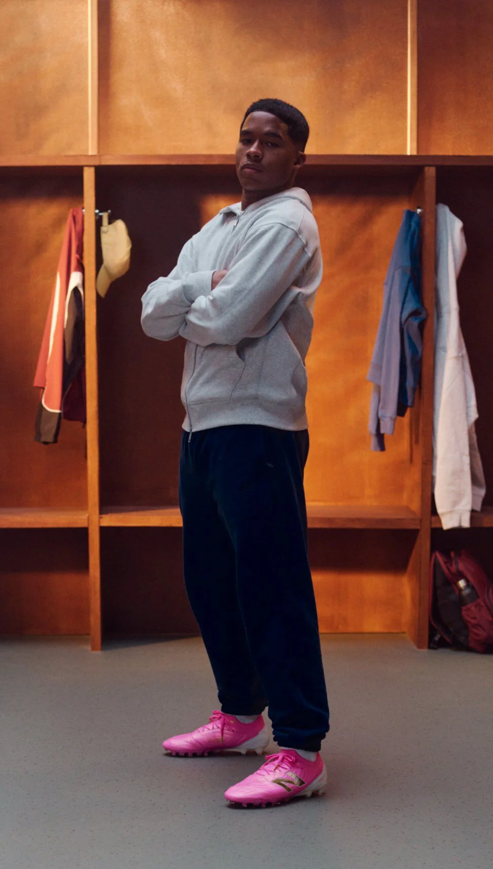 Endrick standing in a wooden locker room with arms crossed, wearing a light gray zip‑up hoodie, dark athletic pants, and bright pink soccer cleats; open lockers behind display hanging apparel, folded clothing, a backpack, and soccer balls on the floor.