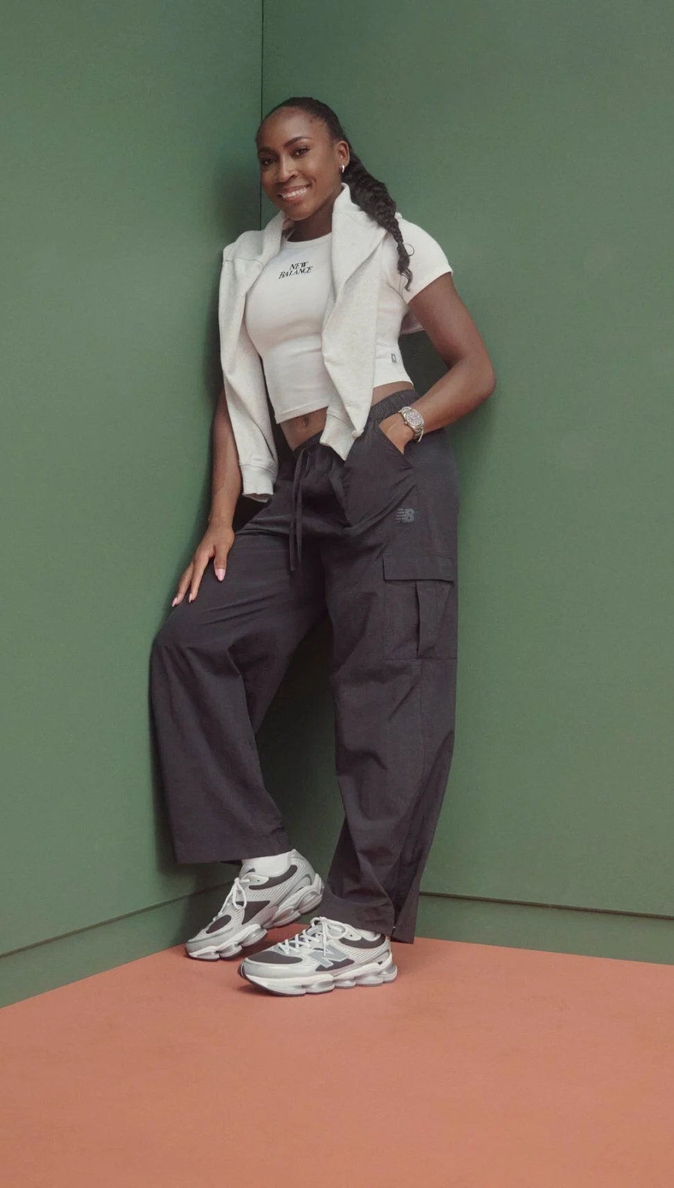 Coco Gauff apoyada en una pared verde en la esquina, con un top corto, pantalones cargo de pierna ancha y zapatillas New Balance blancas y grises.