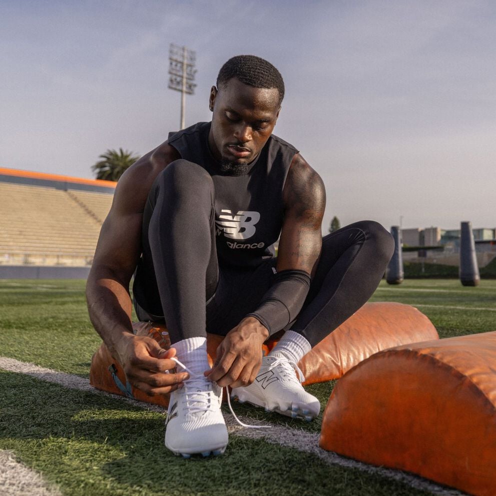 Football athlete in black training gear sits on a football field tying white cleats, with orange padded training equipment nearby.