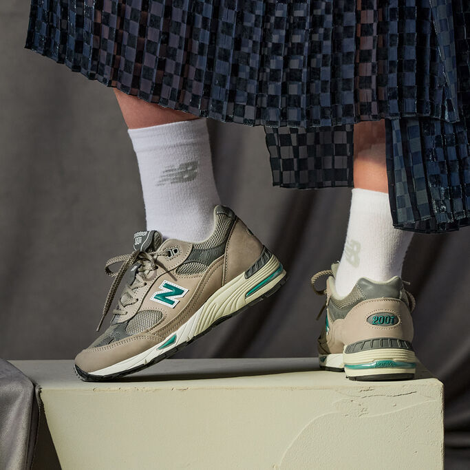 Knee down crop of a woman wearing grey NB 991 sneakers with white crew socks and a long, pleated dark blue skirt