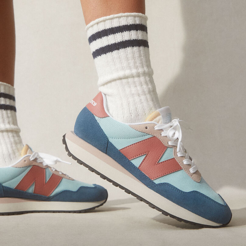 Close up of girl in high socks with NB 237 shoes in blue, teal and salmon posed with toe up in grey studio environment with shadow effects