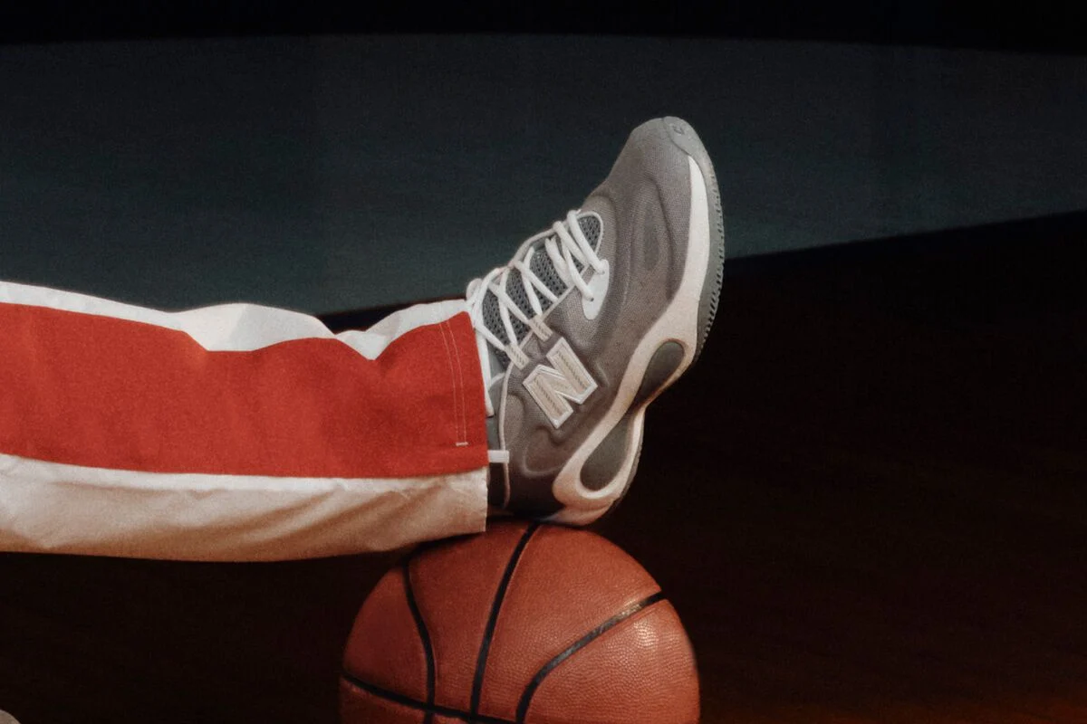 Gray New Balance basketball sneaker resting on a basketball, paired with white and red athletic pants on a dark court.