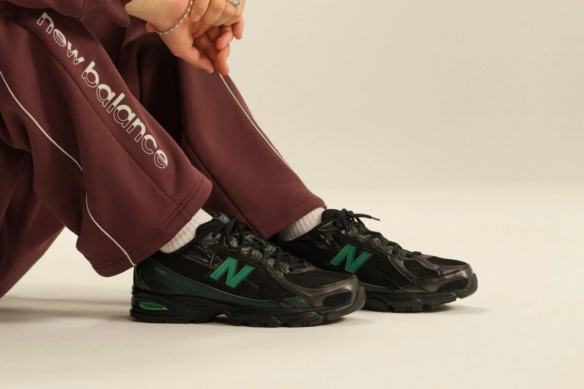 Close-up of black New Balance athletic sneakers with green accents paired with burgundy track pants and white crew socks in a clean studio setting.