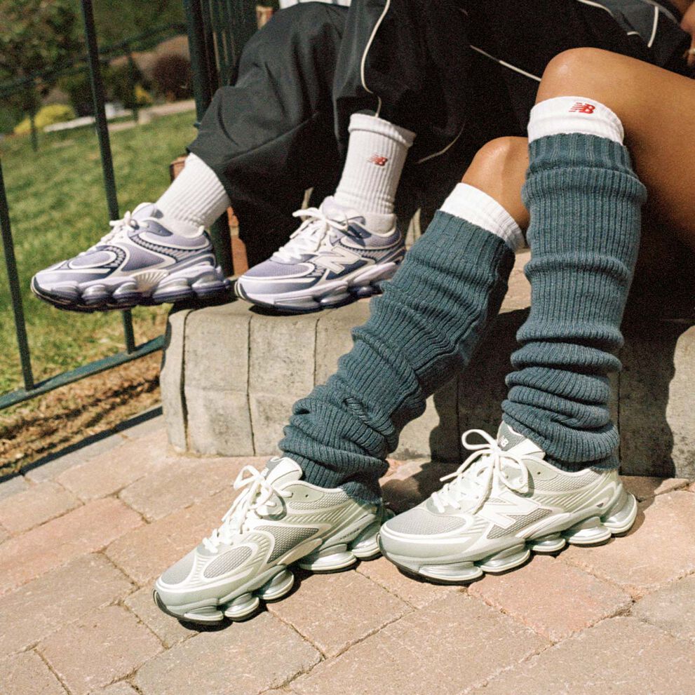 Two people sitting on a sidewalk wearing New Balance sneakers.