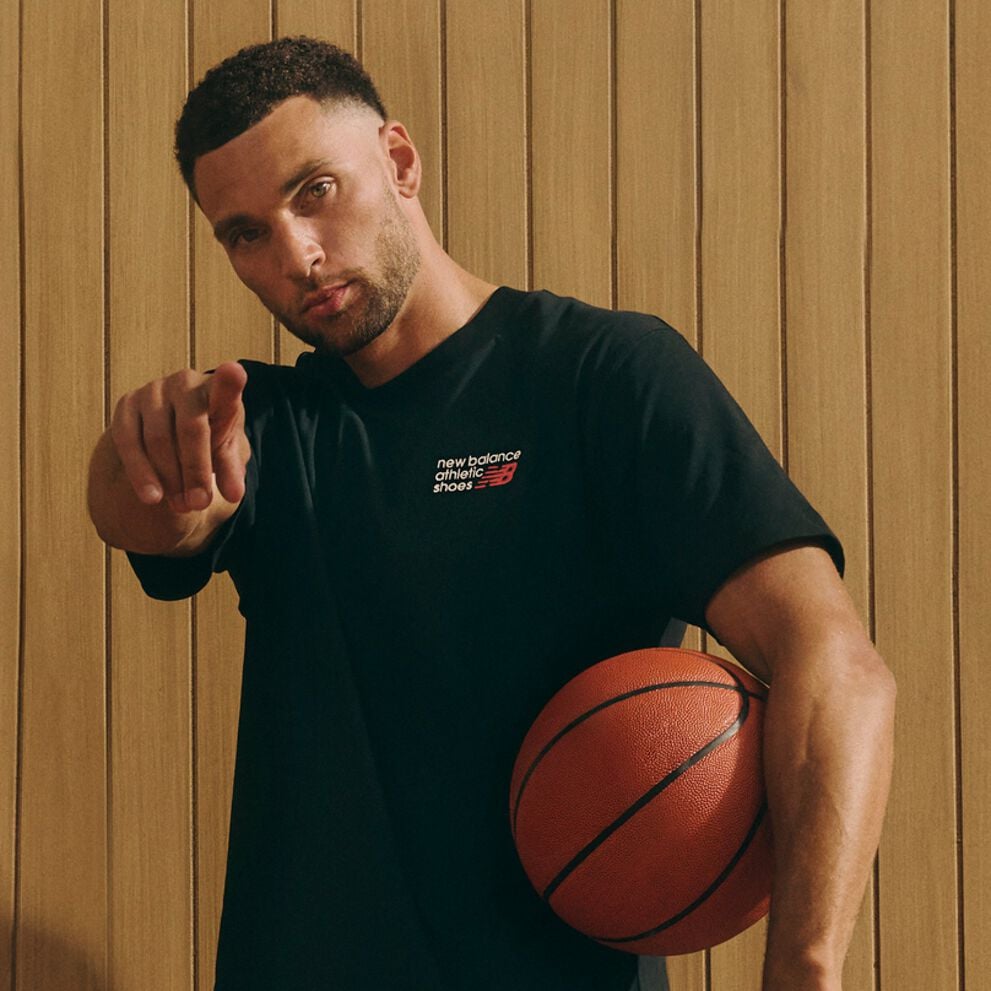 A person in a black New Balance athletic T‑shirt points toward the camera while holding a basketball against their side, standing in front of a wooden-paneled background.