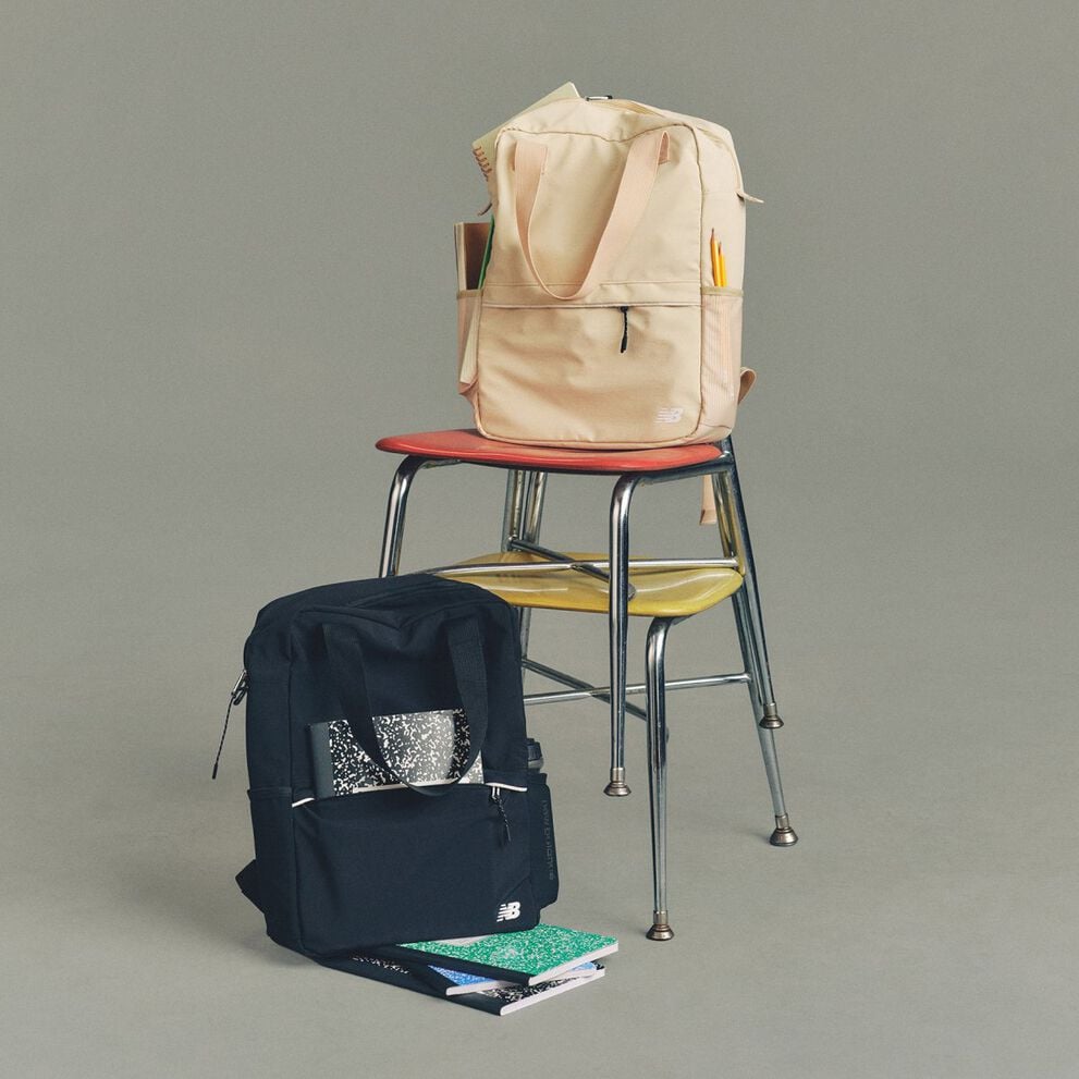 Studio image of a cream colored and a navy colored New Balance backpack placed on a metal chair.