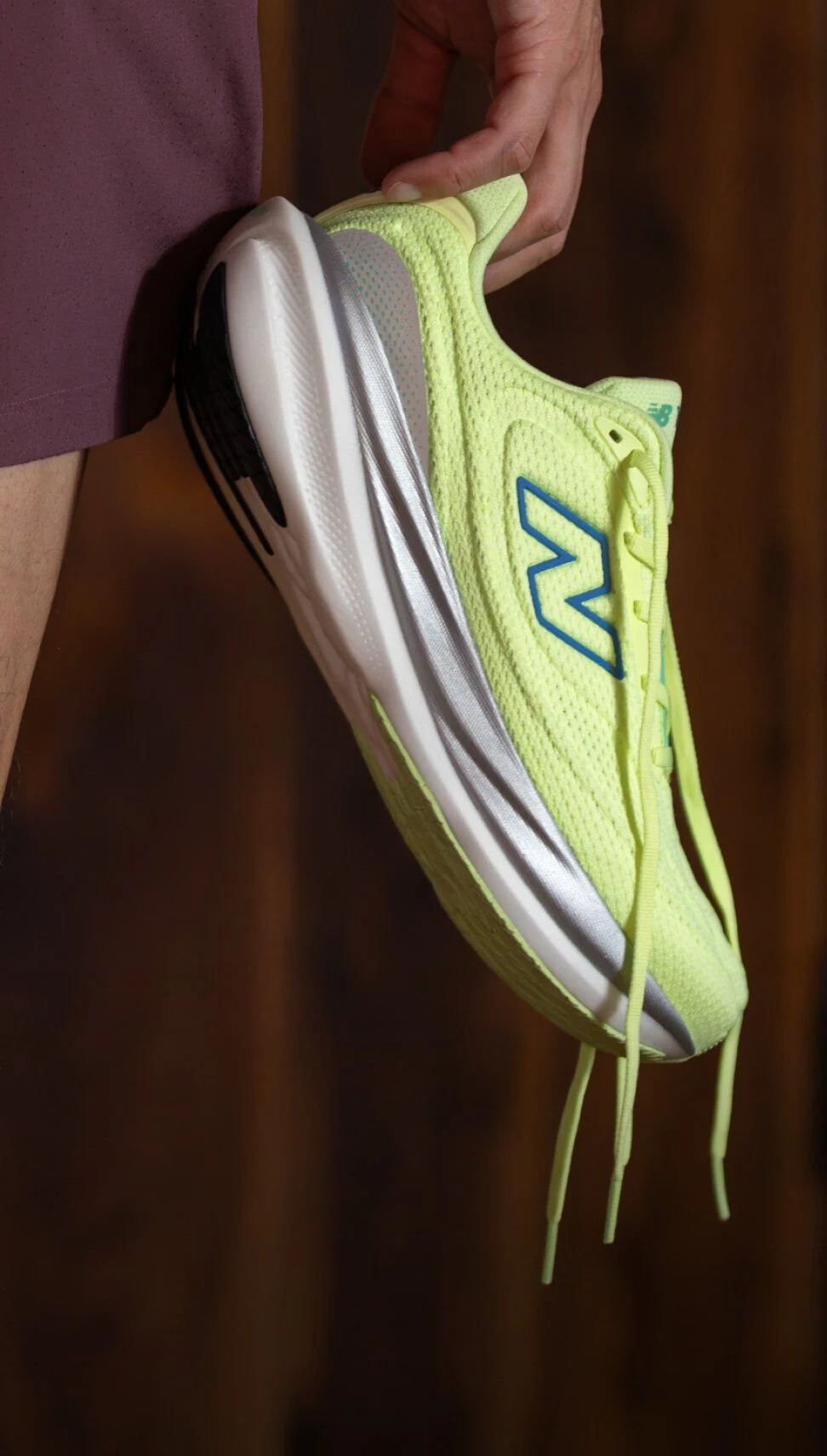 Close-up of a bright yellow-green New Balance running shoe with blue logo and white sole, held by a person wearing maroon shorts. 