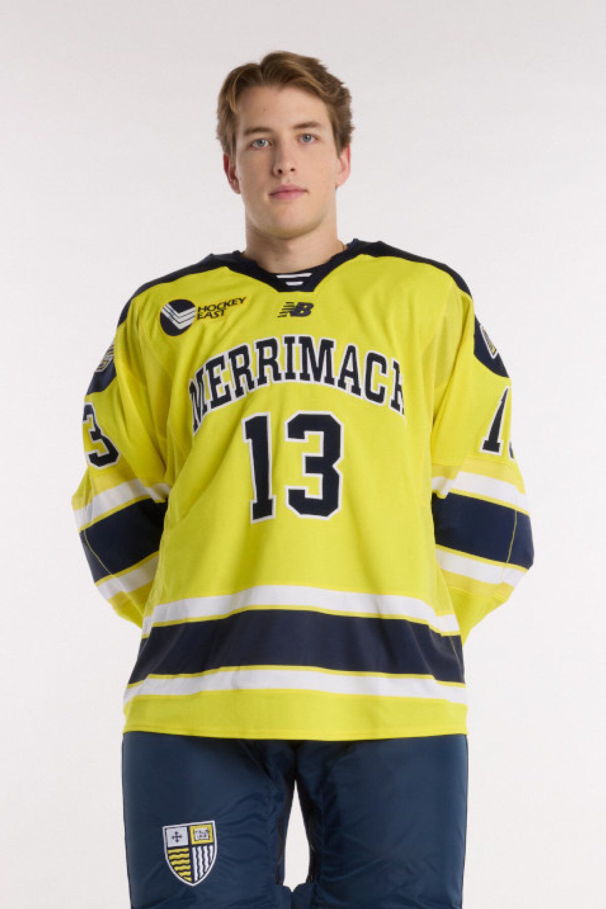 Athlete wearing a yellow Merrimack hockey jersey with number 13 and navy hockey pants, standing against a light background.
