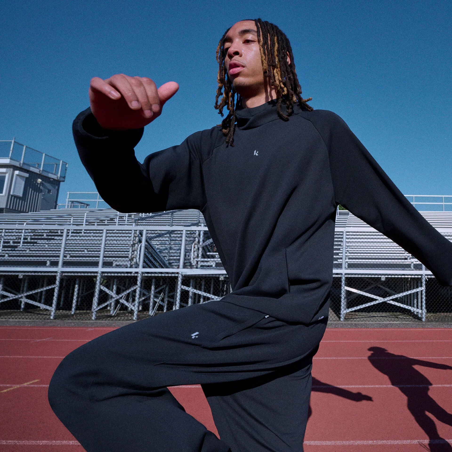 Athlete wearing a black tracksuit performing a dynamic warm-up on a running track in front of stadium bleachers.
