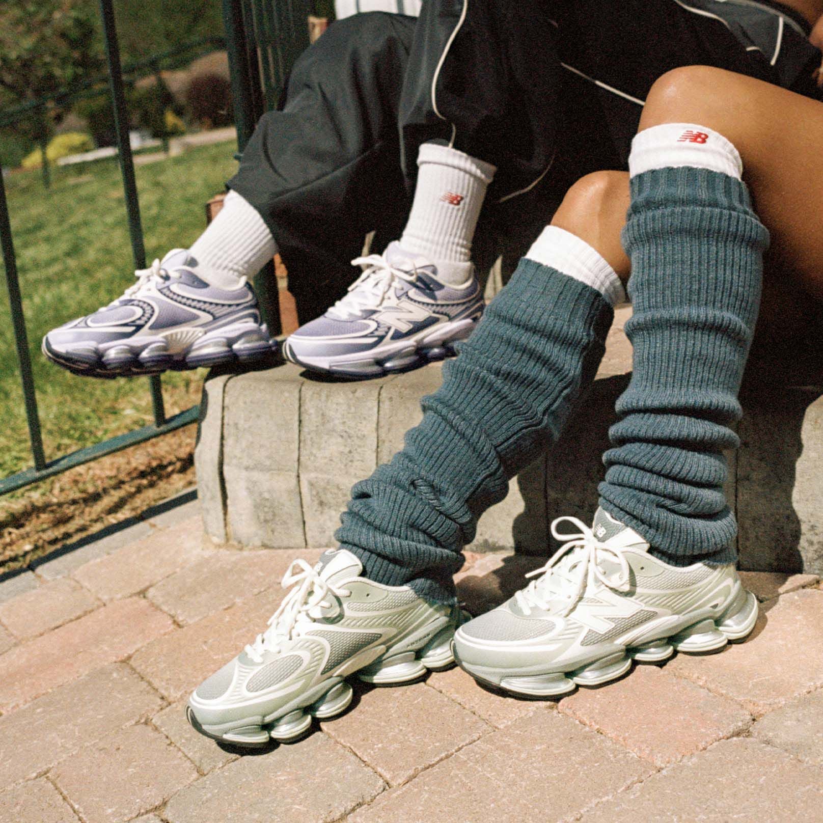 Two people sitting on a sidewalk wearing New Balance sneakers.