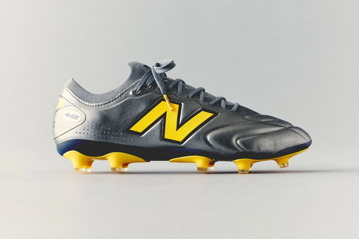 Gray and yellow New Balance soccer cleat with a streamlined synthetic upper and molded studs displayed against a clean white background.